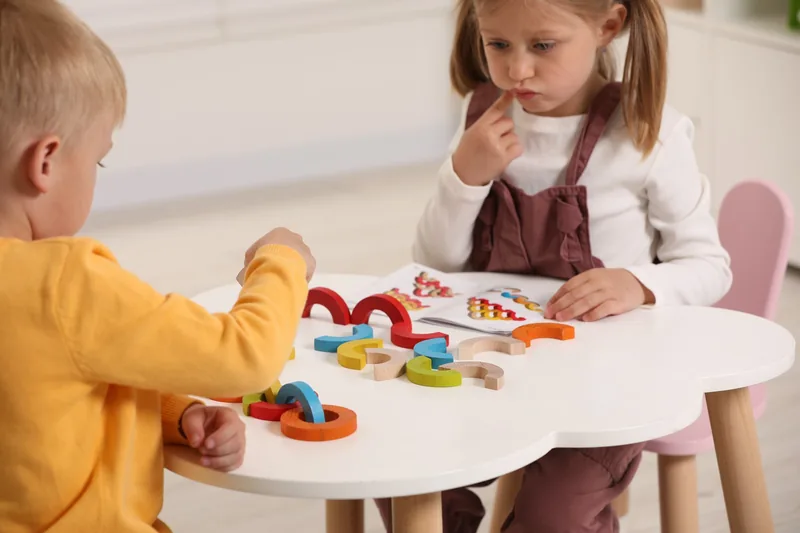 Ein Bub und ein Mädchen sitzen an einem kleinen Tisch und legen aus bunten Holzbögen nach einer Vorlagekarte ein Muster.